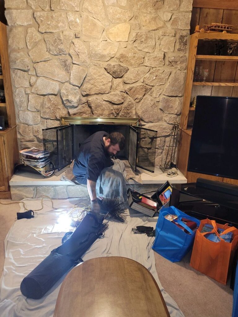 Technician kneeling by a fireplace with chimney sweep tools, preparing for maintenance work in a home's living room.