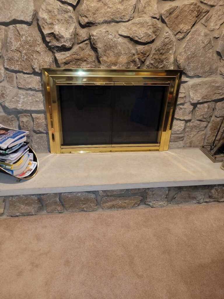 A clean brass-framed fireplace with closed glass doors set in a stone wall, with magazines stacked to the side on the hearth