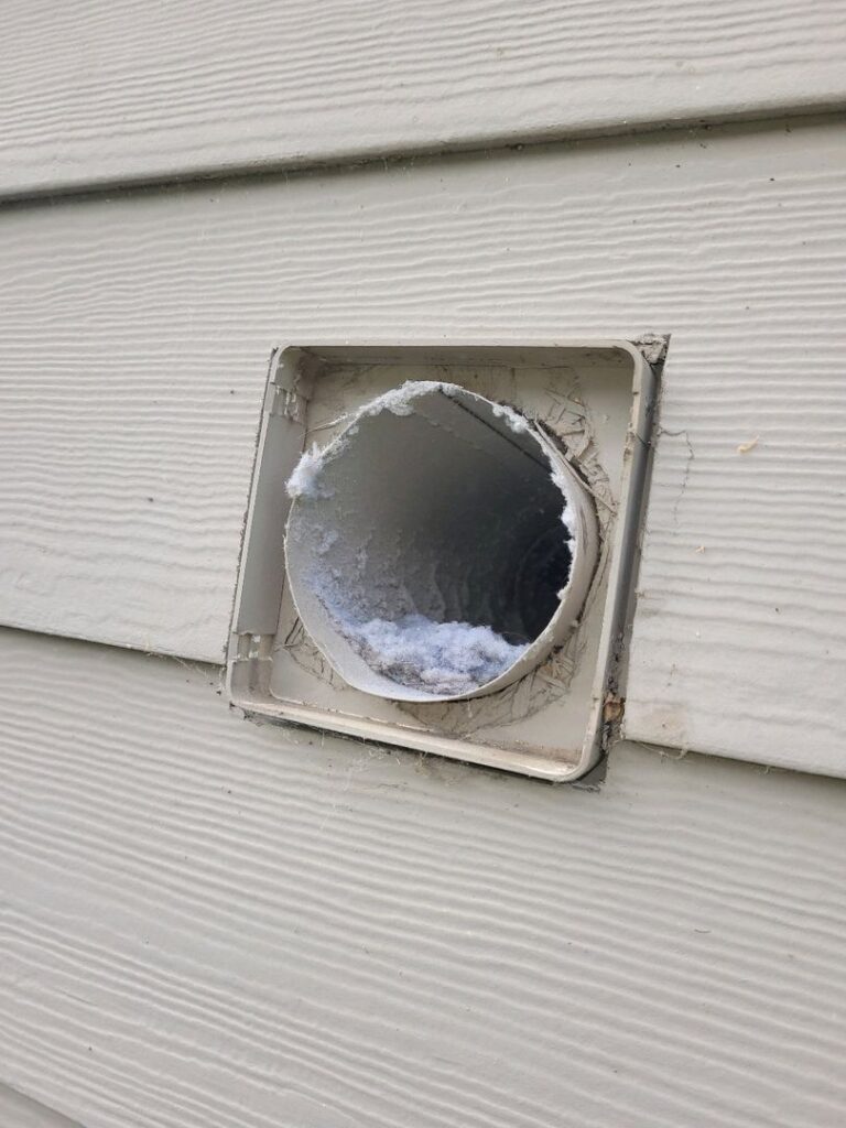 A clogged dryer vent on a house's siding, missing its cover and surrounded by spider webs.