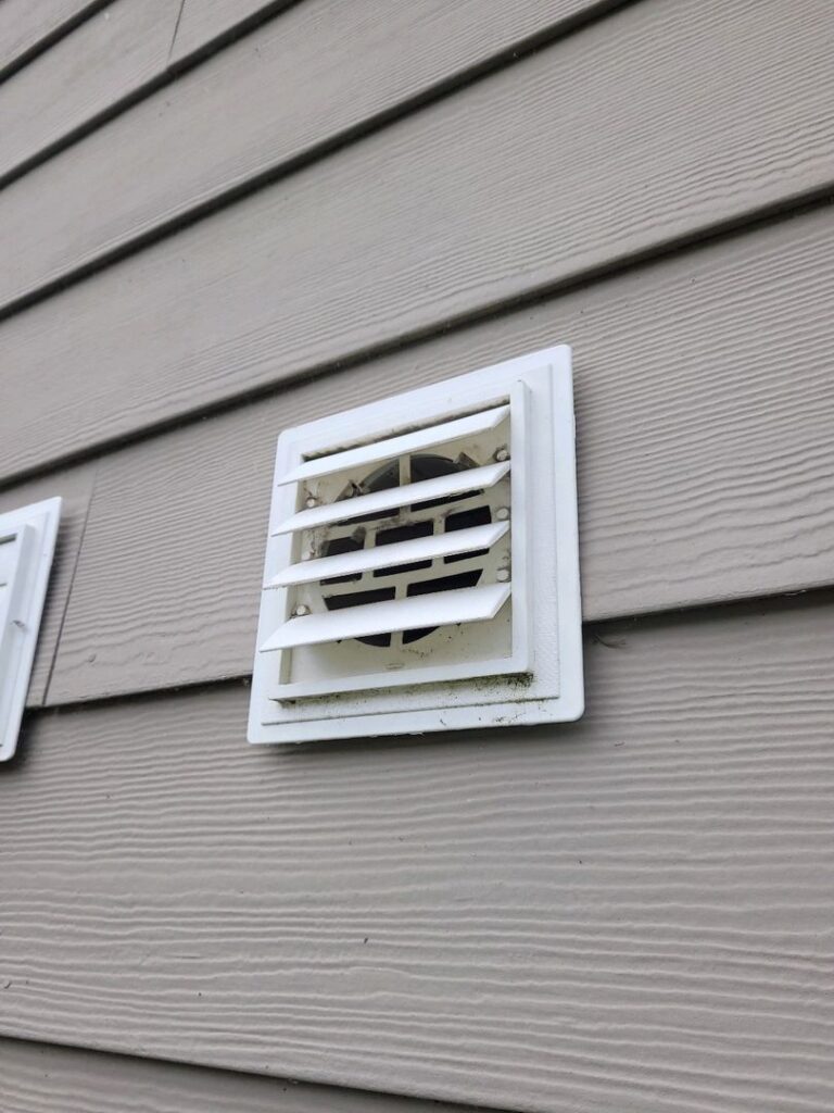 A dirty white dryer vent cover on light-colored house siding