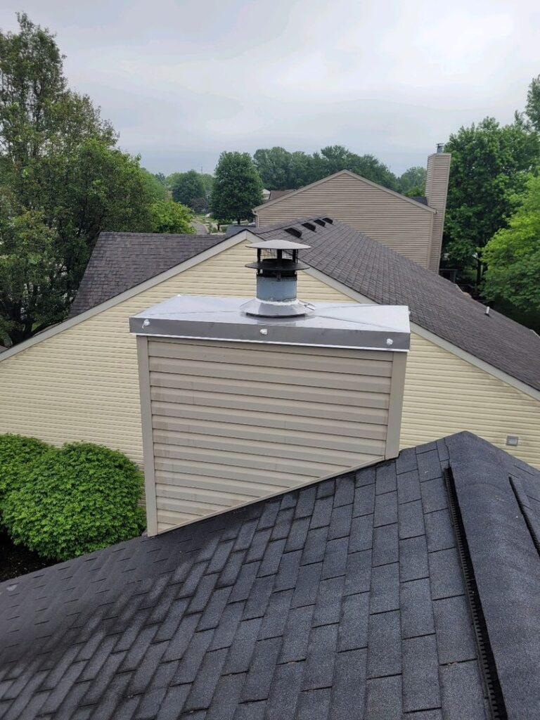 A new stainless steel chase cover on a house chimney with grey shingle roofing and beige vinyl siding.