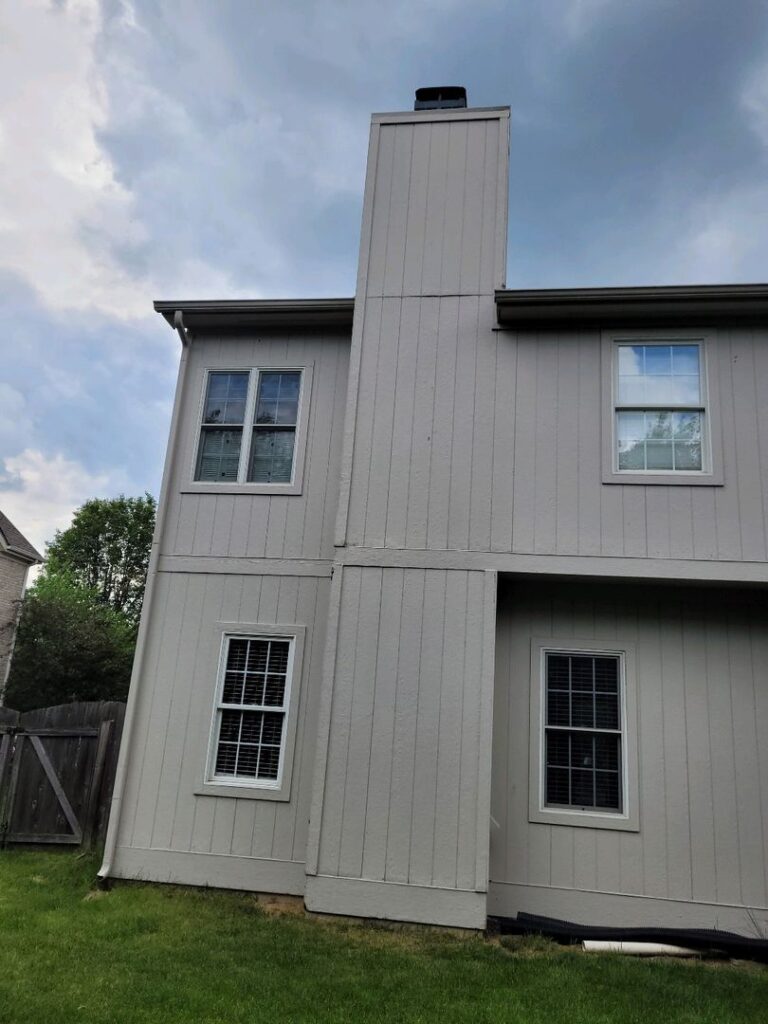 A two-story house with gray siding and a tall chimney, three windows with shutters, and an overcast sky.