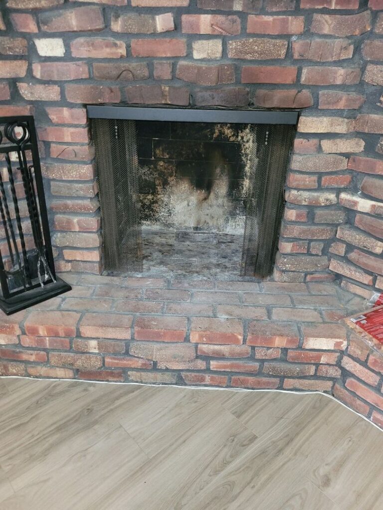 A cleaned out red brick fireplace with soot marks, a black mesh screen, and a matching brick hearth set against light wood laminate flooring