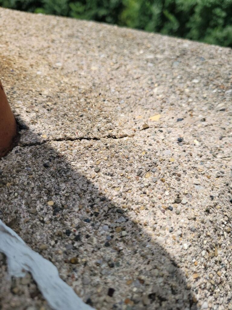 Close-up image showing a crack in a textured concrete chimney crown with out-of-focus greenery in the background