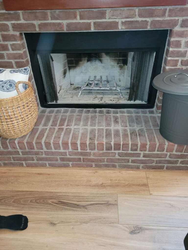 A brick fireplace with metal grate and ash residue, flanked by a wicker basket and metal container, on hardwood flooring.