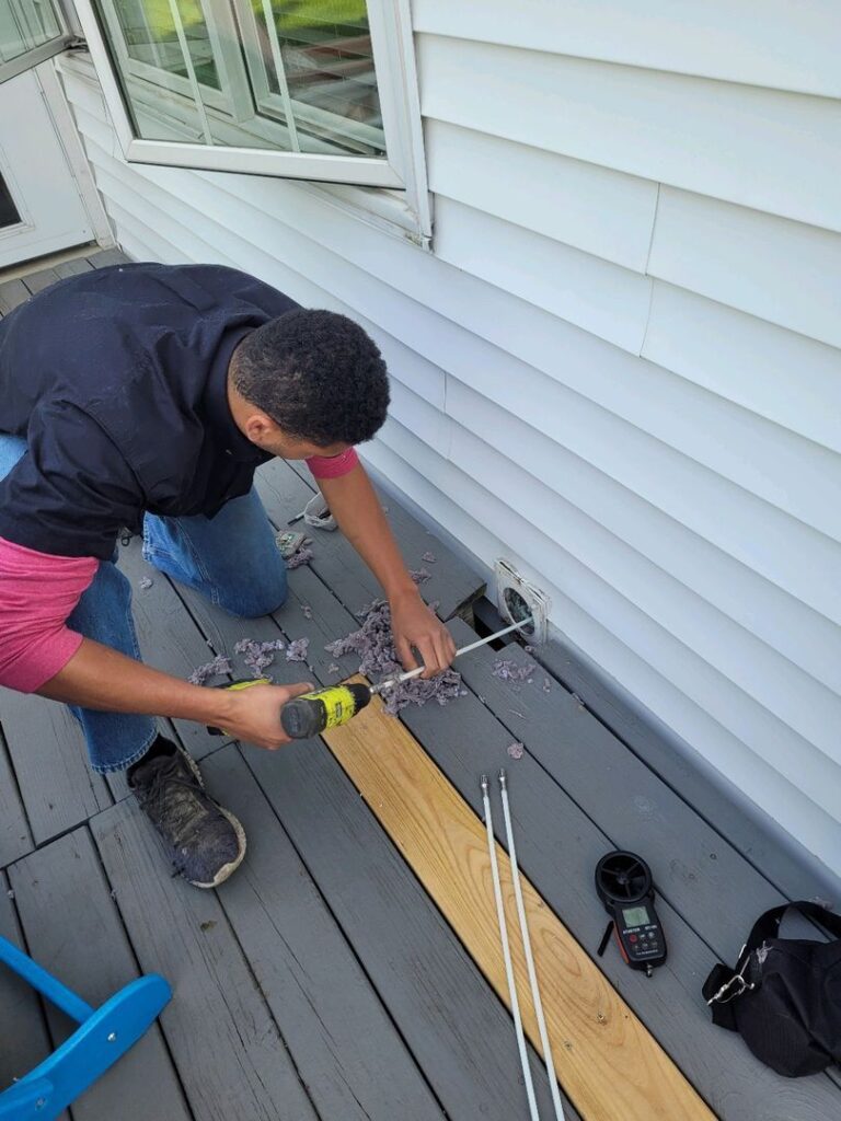 Technician kneeling on a deck while cleaning a dryer vent with a power drill and flexible rods.