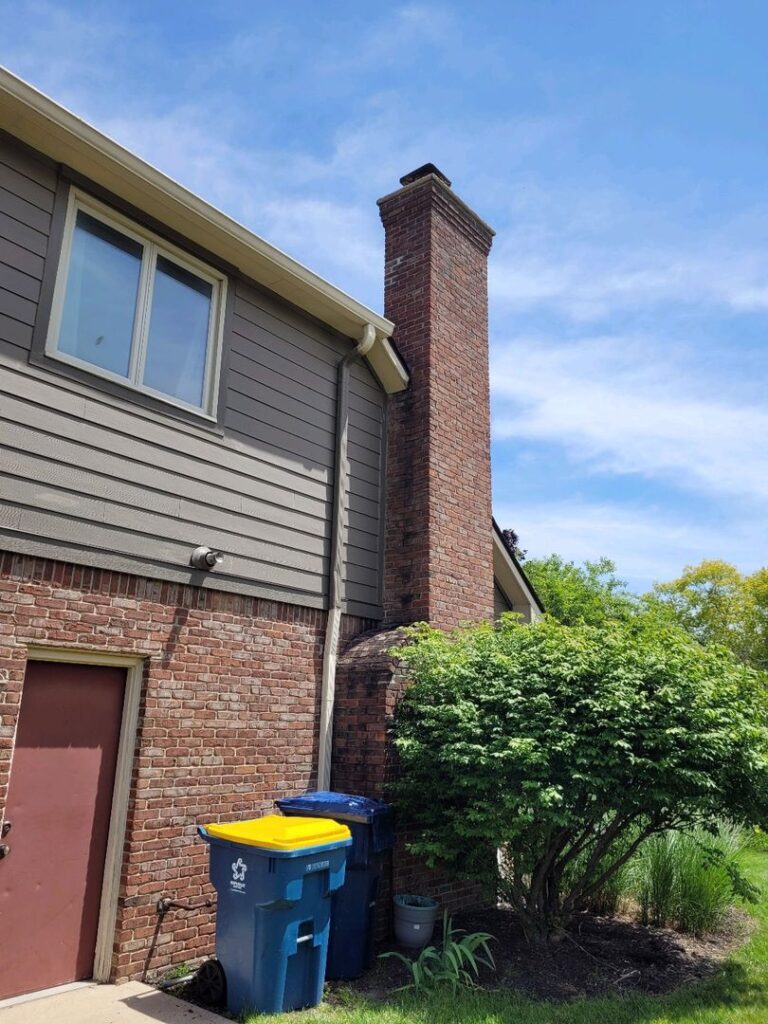 Image of a house with a red brick chimney, grey siding, recycling bins, and green shrubbery under a sunny sky