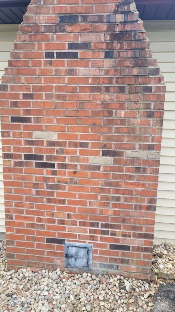 A red brick chimney featuring repair patches and a black metal access door, next to a house with beige siding.