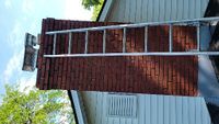 Rotated view of a red brick chimney with a metal cap on a house with white siding, surrounded by greenery and a clear sky.